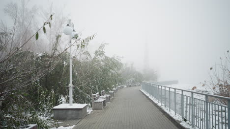 winter cityscape with foggy atmosphere, first snowfall covering park benches, iron railings, light poles, and frosted trees along peaceful walkway