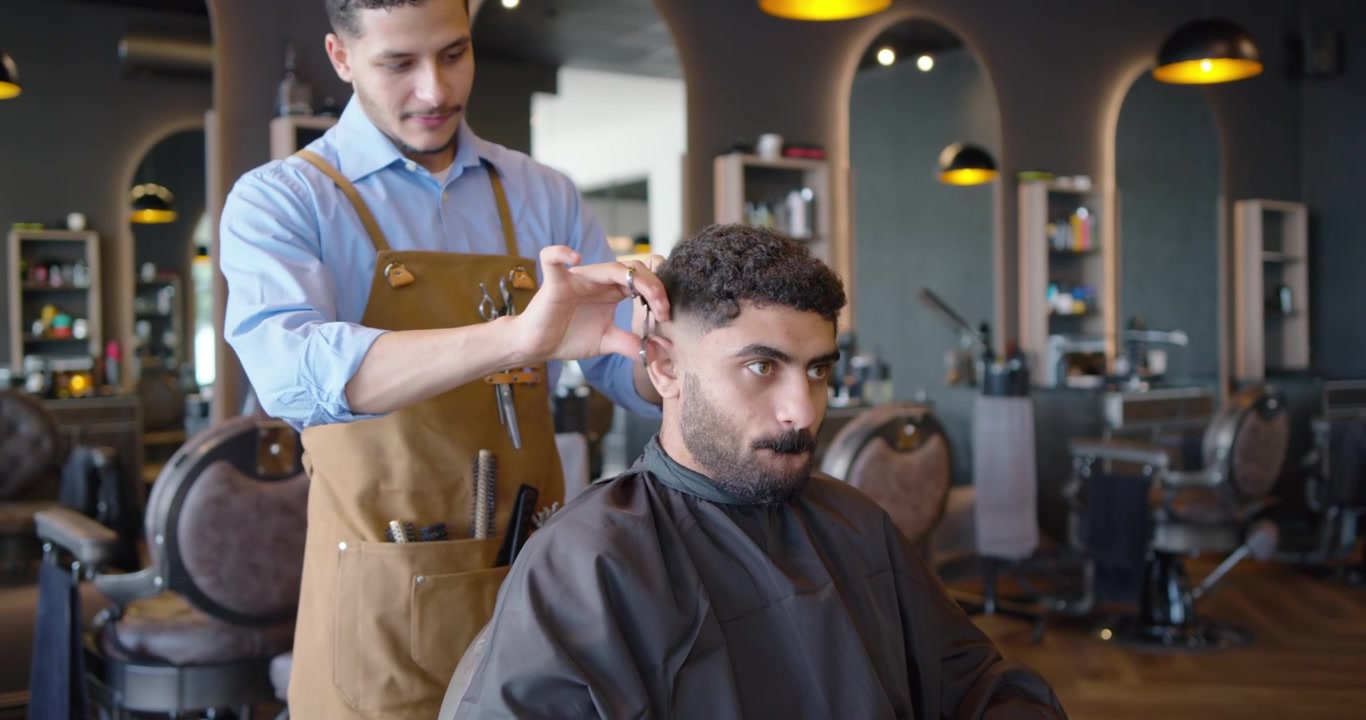 Mandlig frisør klipper en kunde efter at have drejet hovedet, bruger klippemaskine og saks i en moderne salon til en fade-frisure.