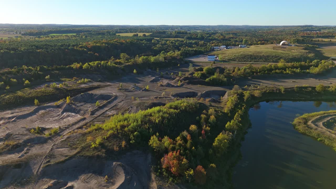 Aerial view of a gravel pit quarry in Caledon showing carved excavation terraces, rugged earth patterns, a reflective water basin, and surrounding autumn foliage across a wide industrial landscape