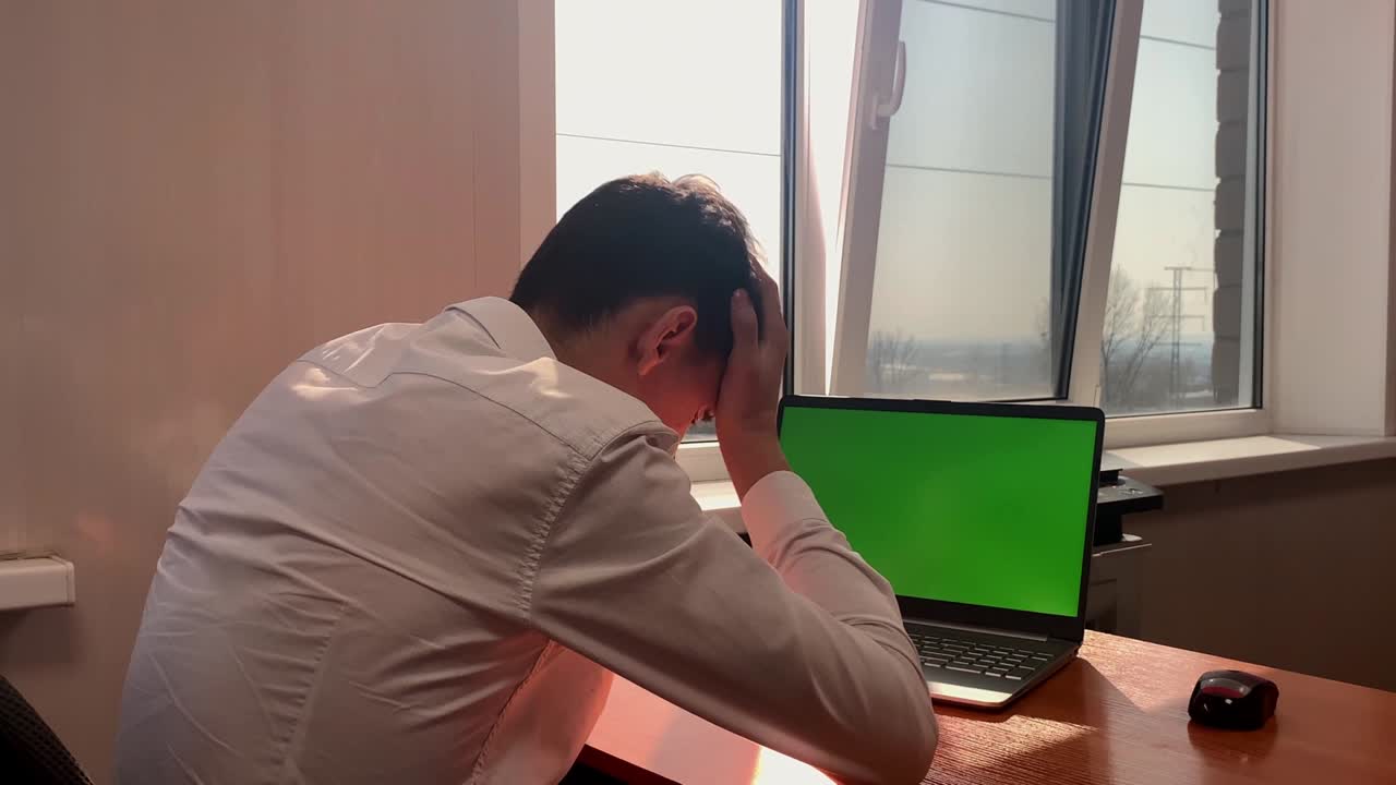 A young man sitting at a computer with a green screen in the office grabs his hair with his hands. Failures at work, fatigue, disappointment