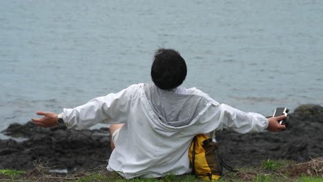 Junge-Fotografin-Genießt-Das-Leben-Und-öffnet-Die-Arme-Zum-Himmel,-Während-Sie-Auf-Der-Klippe-Vor-Dem-Meer-Sitzt