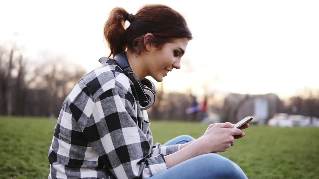 Girl in plaid shirt is sitting on ground in park. She's messaging with a mobile. Happy, smiling. The sun is shining on the background. Side view