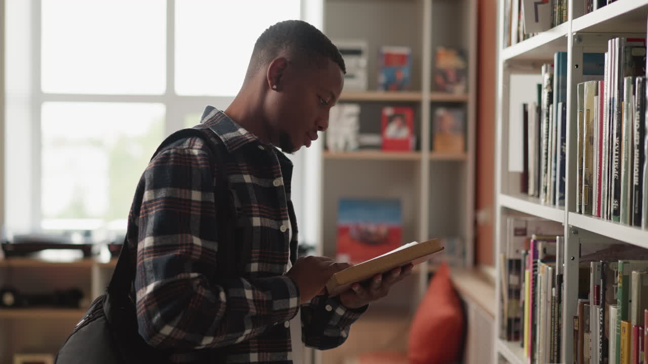 hombre de piel negra lee un libro en la biblioteca. el lector encuentra el libro correcto profundamente inmerso en la lectura se encuentra en la estantería de la librería. el hombre se sumerge en la lectura