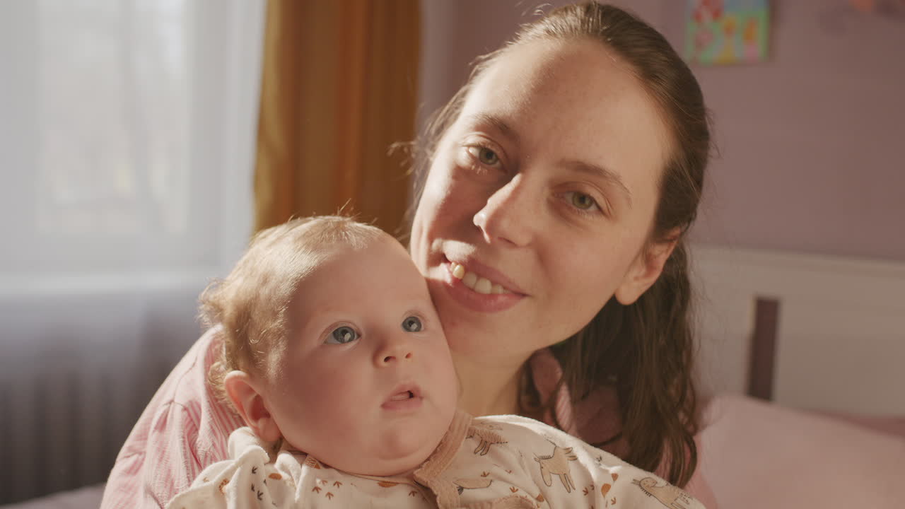 Porträt einer Mutter, die ihr Baby küsst und in einem sonnendurchfluteten Raum in die Kamera lächelt