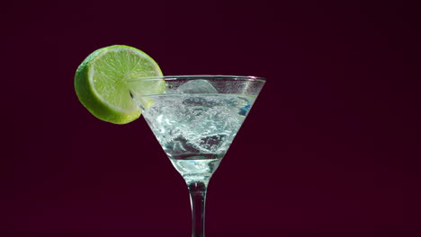 video del producto de un vaso de martini, gin tonic, bebida alcohólica con una rodaja de limón y bloques de hielo, girando en una mesa giratoria con fondo burdeos profundo