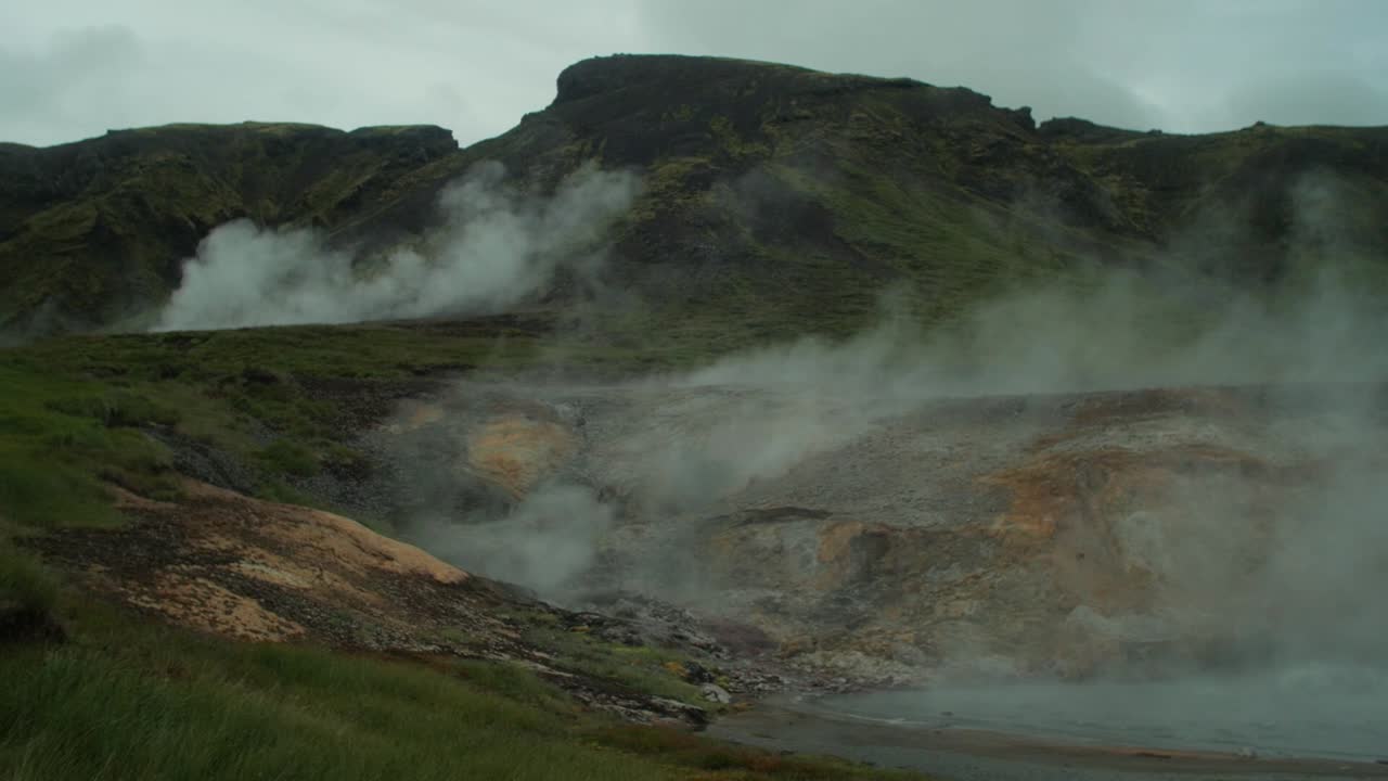 paisaje de islandia, aumento de humo de vapor de aguas termales geotérmicas, figura distante de fotógrafo tomando una foto de una escena completa, lente de gran angular, cámara panorámica de izquierda a derecha
