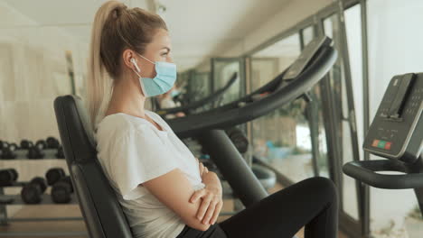 young athlete female with face mask uses an exercise machine and looks to camera