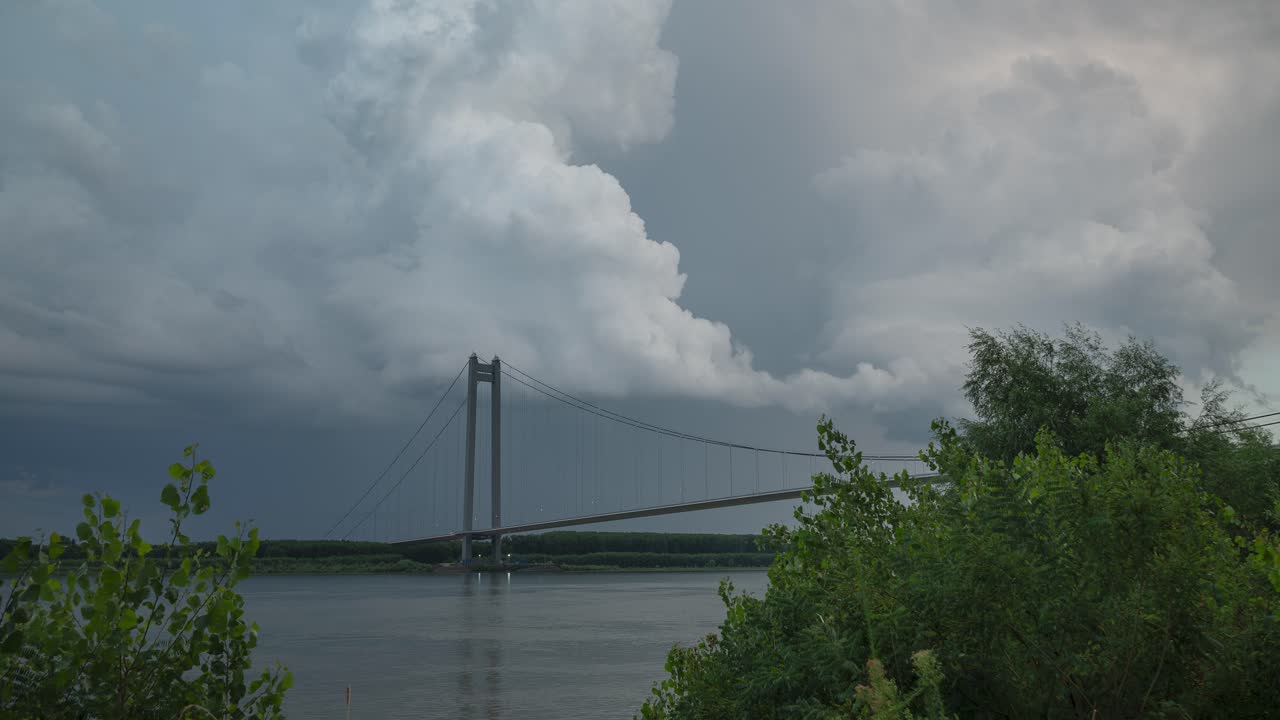 Storm clouds forming above a suspended bridge crossing a big river, vibrant high contrast timelapse