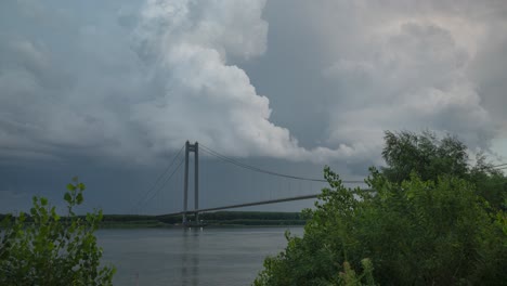 storm clouds forming above a suspended bridge crossing a big river, vibrant high contrast timelapse