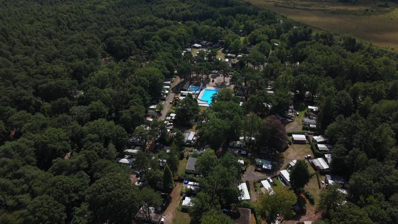 Caravanas e trailers agrupados ao redor de uma piscina azul dentro de um acampamento em floresta densa nos Países Baixos, caminhos de terra serpenteiam entre árvores imponentes sob a luz quente do sol de verão. Tomada aérea ampla de drone