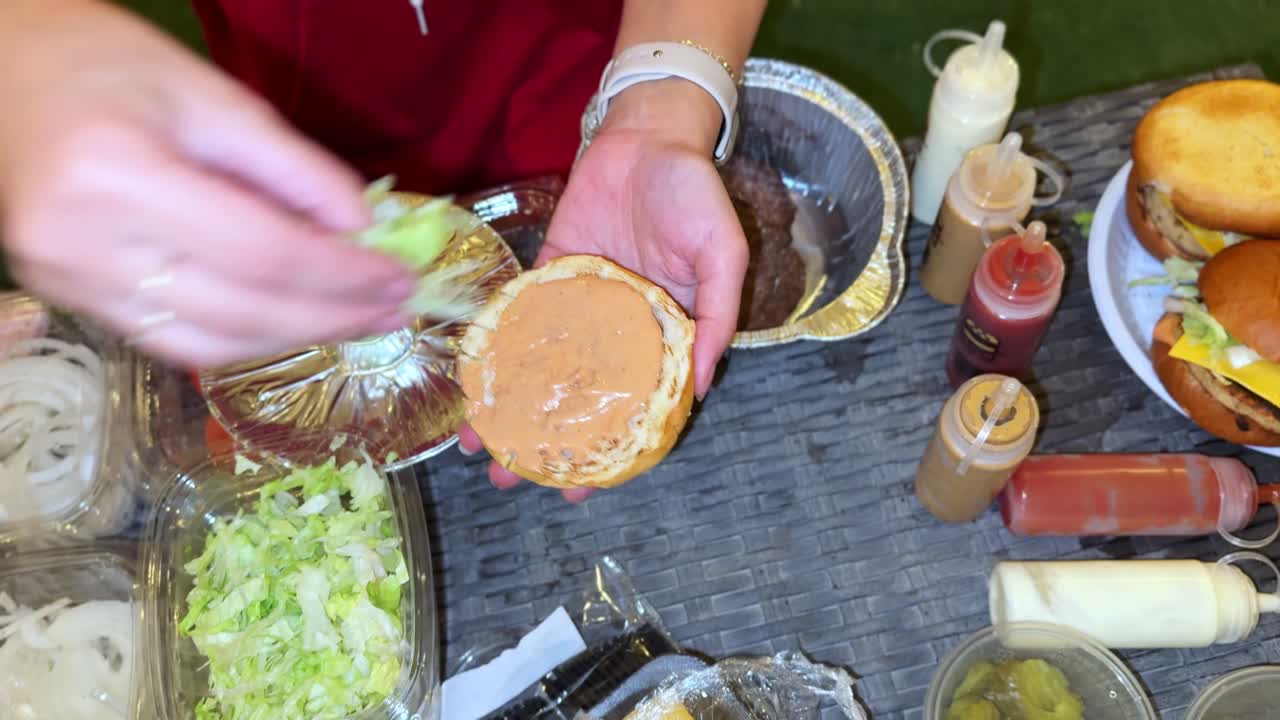 Hands preparing a homemade burger with fresh lettuce, tomato, cheese, sauce and a juicy patty. Close-up cooking scene showing delicious step-by-step burger assembly