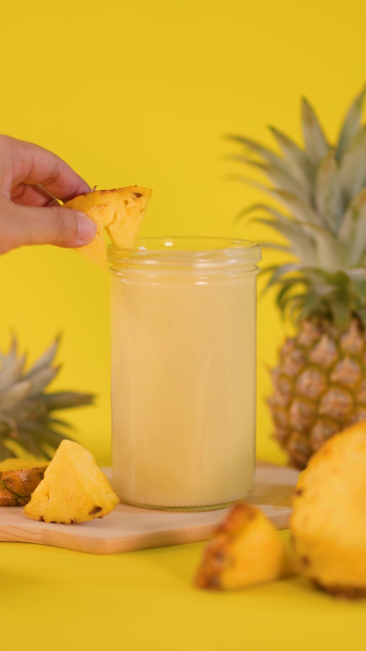 Una mano posiziona una fetta d'ananas su un frullato cremoso, circondato da ananas, con uno sfondo giallo brillante e illuminazione soffusa.