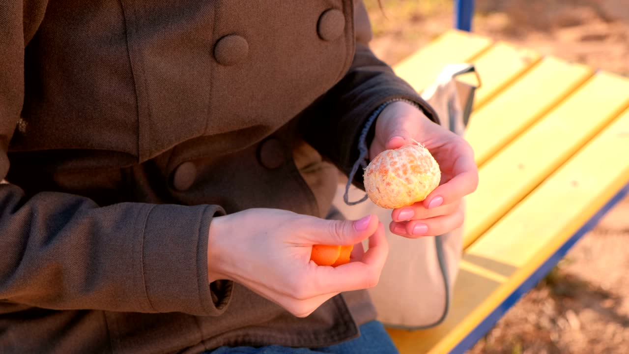 선글라스를 입은 젊은 갈색 여성이 공원의 벤치에 앉아 망가린을 벗니다. 가까운 손.