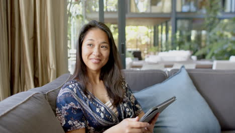 happy asian woman using tablet and looking out window in sunny living room, slow motion