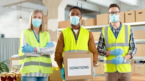 grupo multiétnico de trabajadores de caridad jóvenes y mayores con máscaras faciales que sostienen una caja de donación y miran la cámara en un almacén de caridad