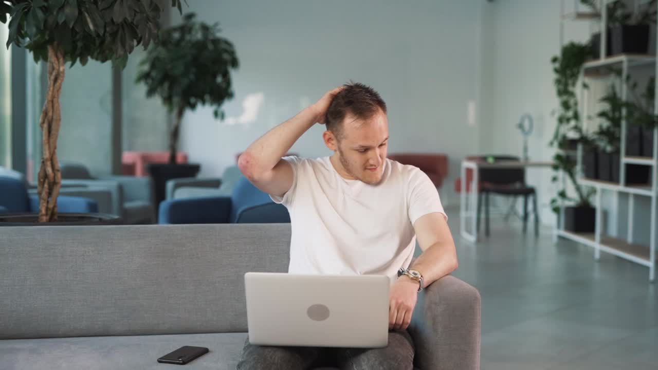 hombre joven en el vestíbulo del centro de negocios está sentado con una computadora portátil, buscando confundido y molesto