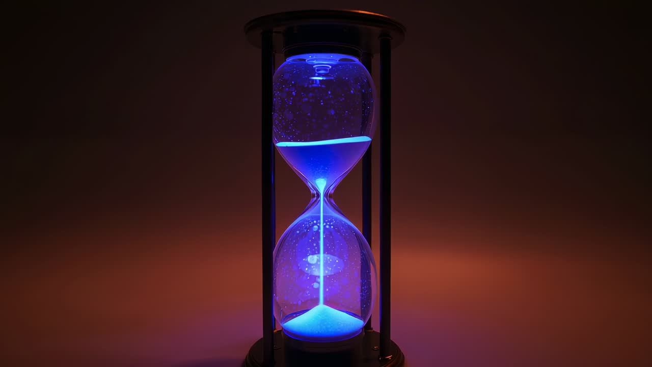 Gravity driving framed hourglass trickling glowing blue sand through neck in studio, measuring time