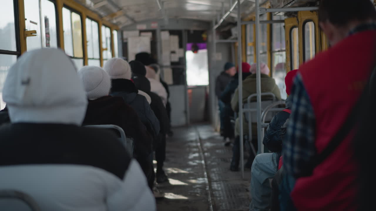 bundled passengers seated inside moving bus, transparent windows reveal bright winter street, soft sunlight and reflections across metal seats and aisle create calm urban commute mood, steady ride