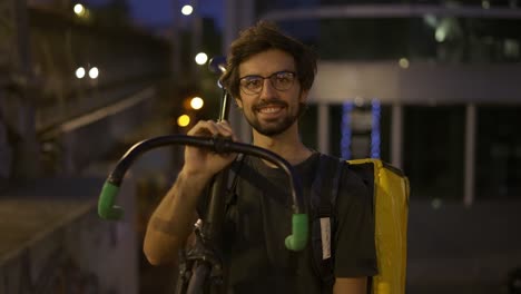 Courier-holding-his-bike-and-backpack-outdoors-and-smiling