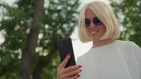 stylish woman in white polo with dark sunglasses flings hair while filming playful moment with phone, showing excitement on face, green trees background, casual outdoor summer scene