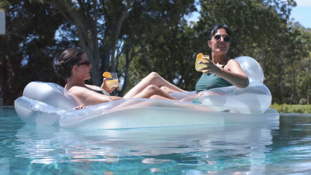 Diverse Freundinnen treiben auf einer transparenten Liege im Pool, eine Frau rechts hebt einen Cocktail und unterhält sich.