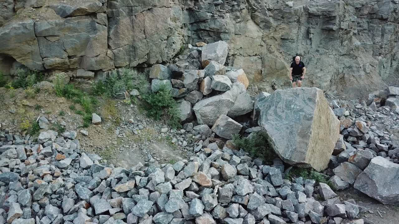 Happy male tourist climbing on the rocky mountains. Active hiker in casual clothes walking through the stones coming into the biggest one and raises his hands up.