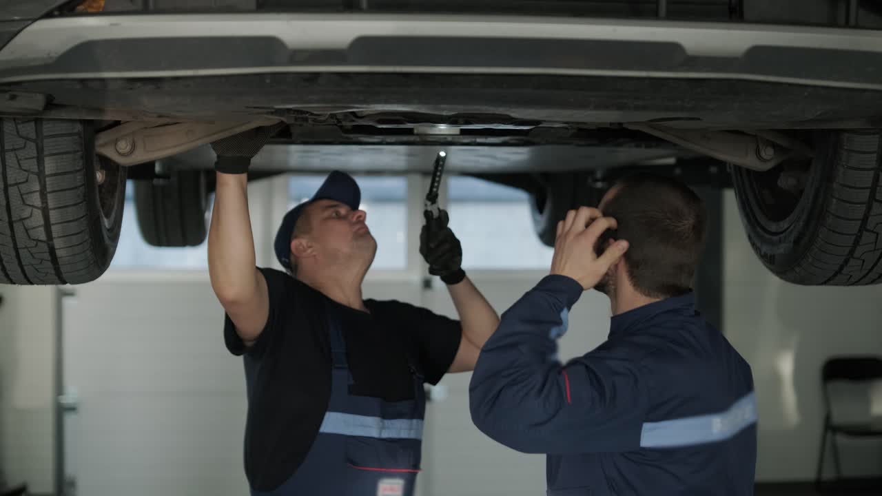 mecánico de automóviles con uniforme azul inspeccionando y reparando la parte inferior de un vehículo levantado en un taller de reparación profesional. el taller está bien iluminado y equipado con herramientas
