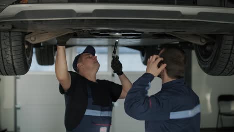 auto mechanic in a blue uniform inspecting and repairing the underside of a lifted vehicle in a professional repair shop. the workshop is well-lit and equipped with tools