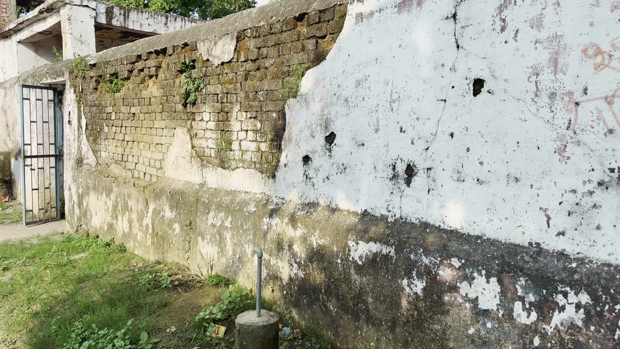A small metal water pipe emerges from a concrete base beside an old weathered wall in a rural area. The scene captures the simplicity of local infrastructure and everyday village surroundings