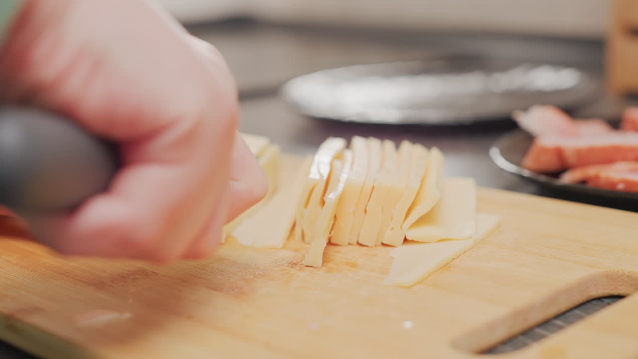 extreme close up of hand using sharp knife to slice yellow dairy product into neat thin strips on wooden cutting board, with blurred kitchen elements in background