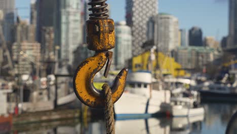 Rotating-close-up-of-yellow-metal-industrial-chain-and-hook-to-lift-cargo-anchor-and-secure-boats-in-the-marina-with-a-blurry-cityscape-in-the-background-blue-sky-buildings-boats