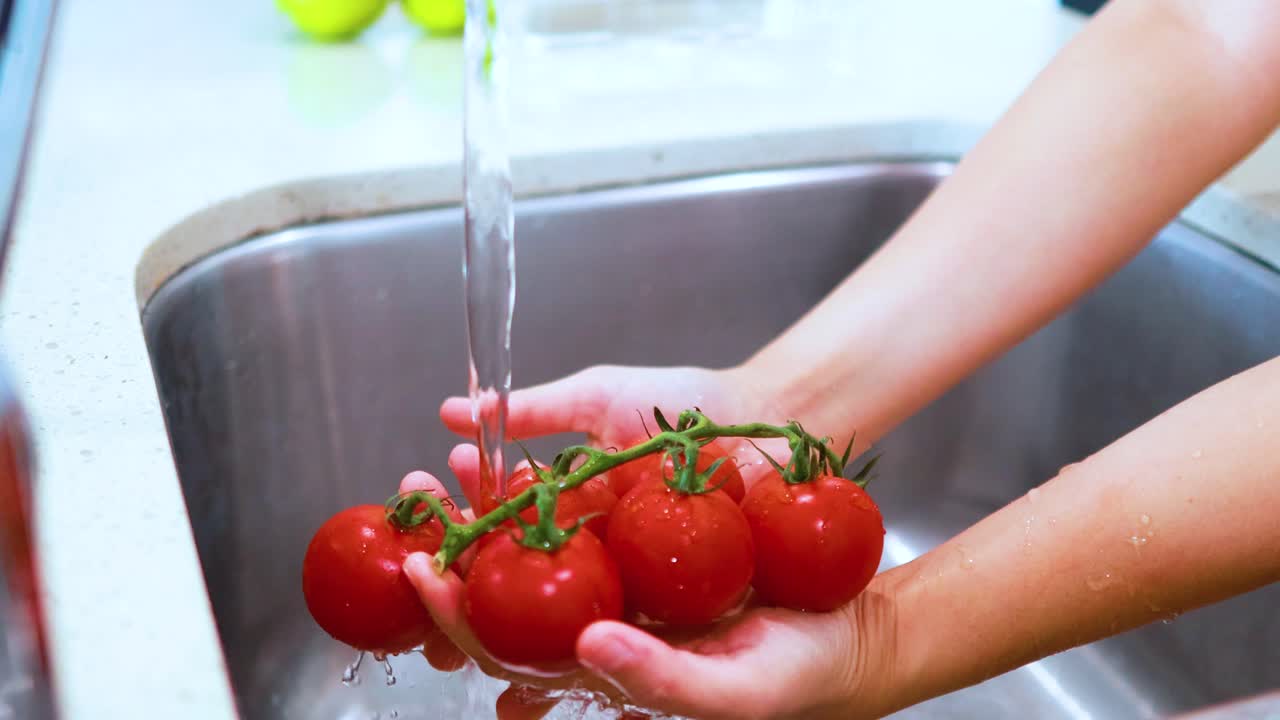 Hands rinse a cluster of ripe vine tomatoes under running water in a modern kitchen sink, with bright natural lighting and steady camera framing