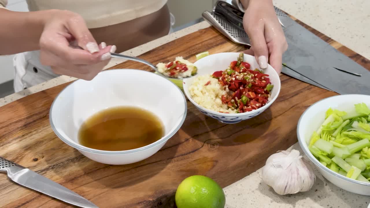 A person prepares a Thai dipping sauce by spooning chopped chili and garlic into a bowl of brown liquid, on a wooden kitchen counter under bright lighting