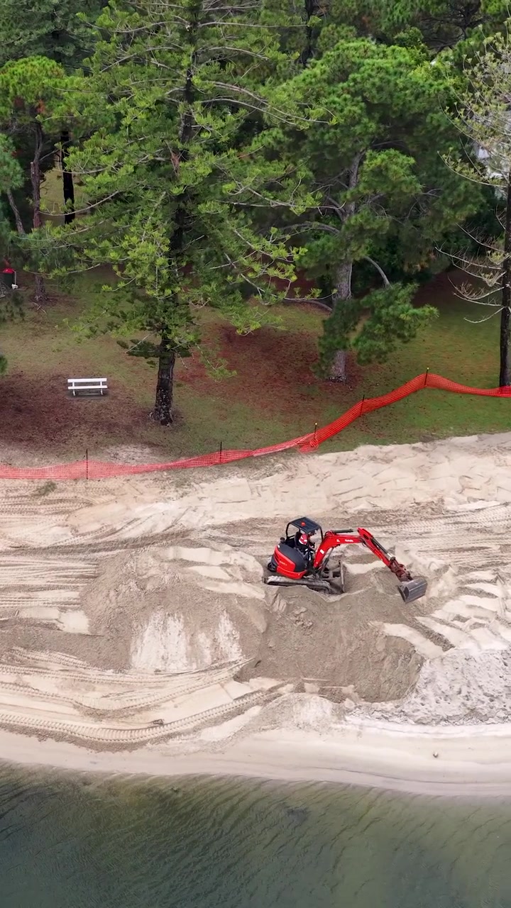 Inquadratura dall'alto di un escavatore rosso che sposta sabbia per la ristrutturazione della spiaggia vicino a case sul lungomare.
