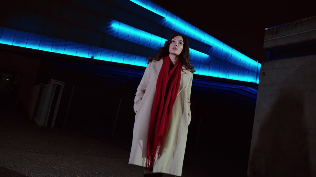 Wide-angle cinematic shot of a woman walking at night beneath blue neon lighting, wearing a coat and red scarf in a modern urban setting. Concept for nightlife, independence, street fashion, city