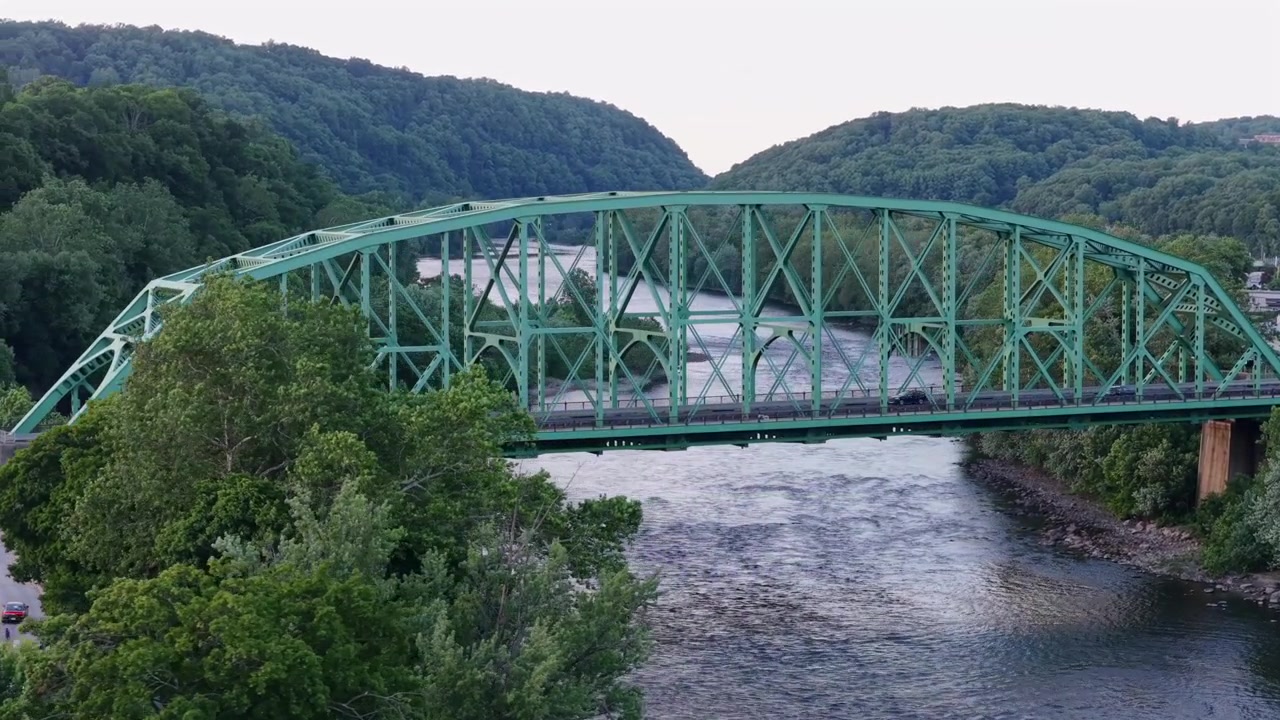 Arched green steel truss bridge spans winding river near Easton Pennsylvania, dense forested hills rise above rocky banks near distant riverside industrial buildings. Aerial drone approaching shot