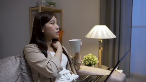 portrait of an asian female manager holding coffee is touching her neck while working overtime pondering on a business strategy in the living room at home