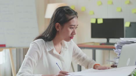 사무실에서 문서와 함께 작업하는 동안 노트북을 보고 메모를 취하는 아시아 여성의 클로즈업