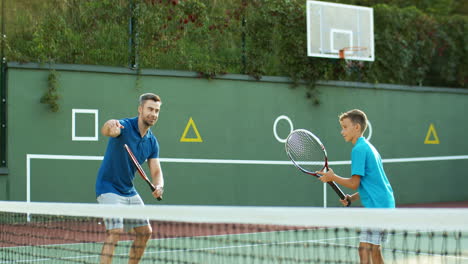 Man-Teaching-His-Teen-Son-How-To-Play-Tennis-On-An-Outdoor-Court-In-Summer