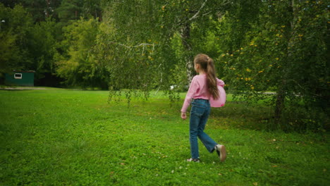 back view of girl child in pink top and jeans walking across grassy field holding balloon under arm, with hair flowing and trees surrounding in peaceful green outdoor park on calm daylight