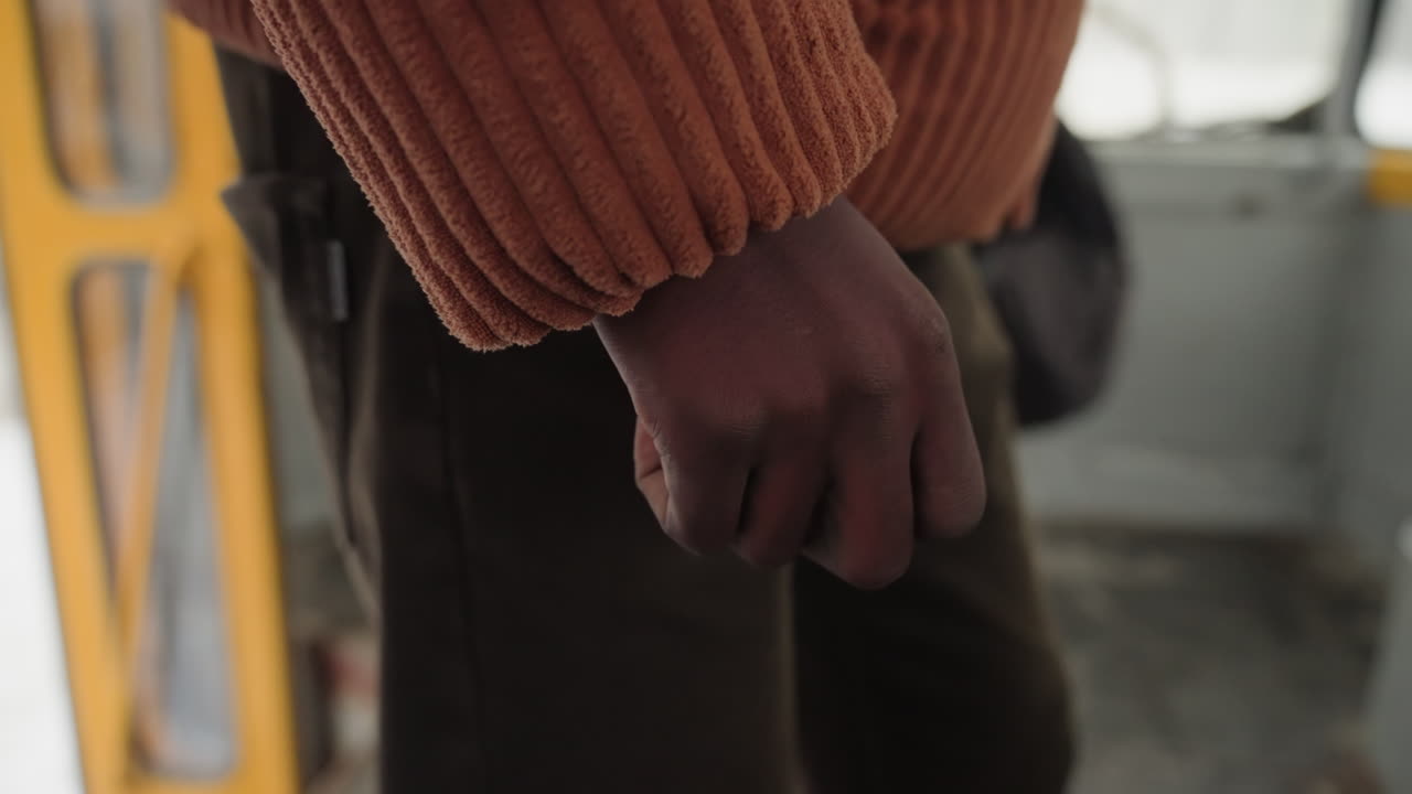 Primo piano della mano di un pendolare vicino alla porta del tram, con un passante che si avvicina sullo sfondo, movimento rapido e leggera sfocatura, manica a maglia e presa salda sul telaio, strada fredda visibile oltre l'ingresso, tensione.
