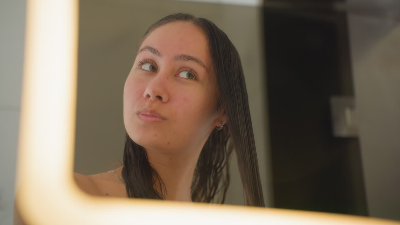 Specchio del bagno dell'hotel, ospite che si spazzola i capelli, luce LED calda e soffusa, capelli umidi e intrecciati, asciugamano drappeggiato sulle spalle, sorriso sottile, riflesso ravvicinato, routine di cura personale, viaggiatore che si prepara