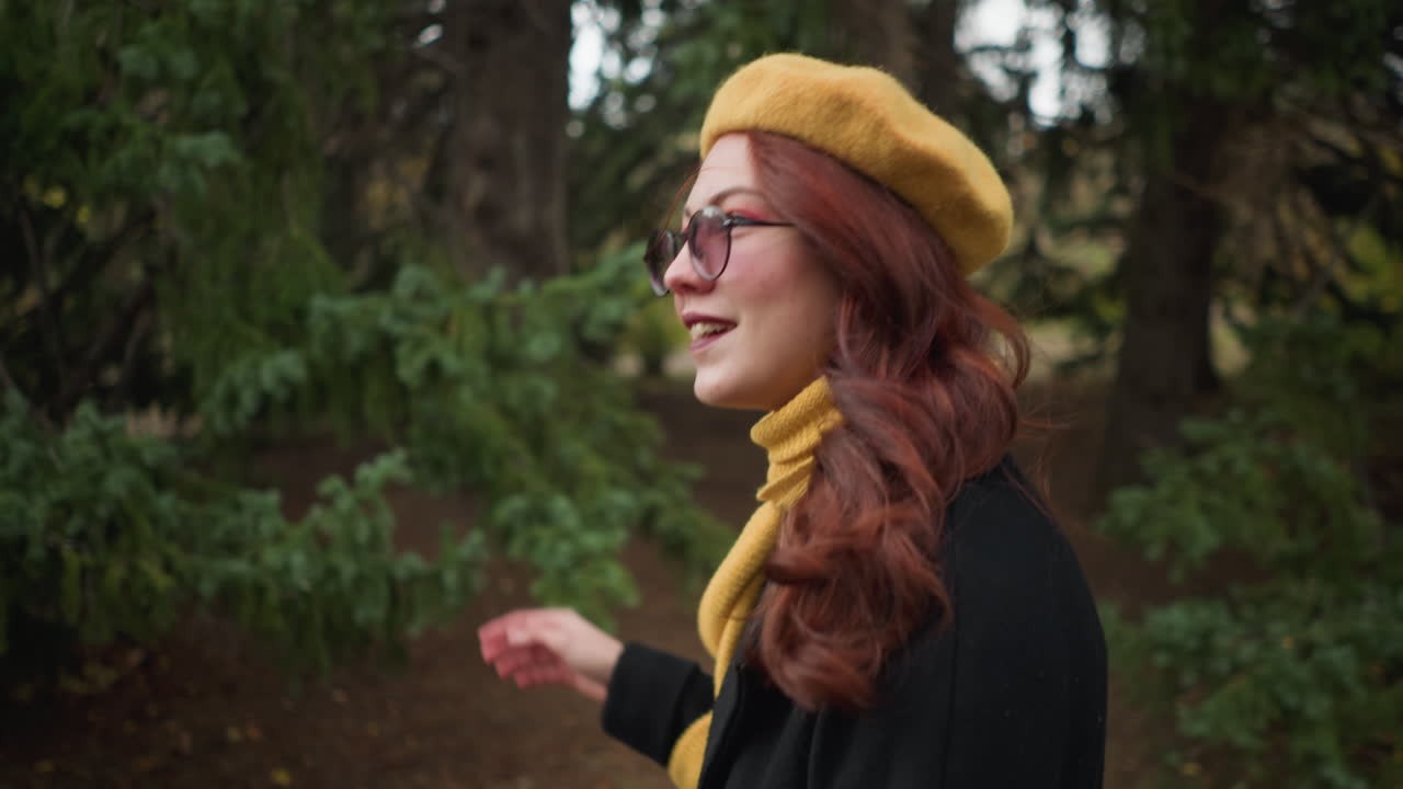 College lady with long wavy red hair, wearing a black coat and yellow beret with matching scarf, adjusts her sunglasses while looking up, walking through a green park with colorful autumn foliage