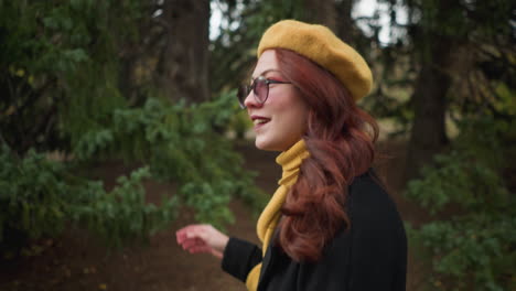 dama de la universidad con largo cabello rojo ondulado, con un abrigo negro y boina amarilla con pañuelo a juego, ajusta sus gafas de sol mientras mira hacia arriba, caminando a través de un parque verde con colorido follaje de otoño