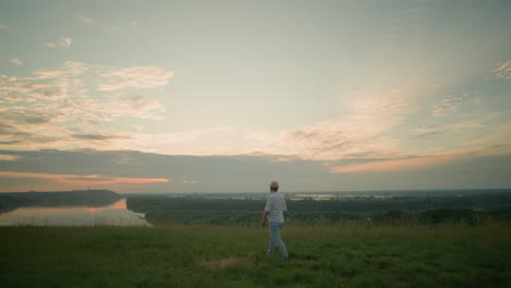 a man dressed in a white shirt, hat, and jeans walks through a grassy field towards a serene lake at sunset. he pauses with his arms folded, contemplating the peaceful landscape and tranquil waters