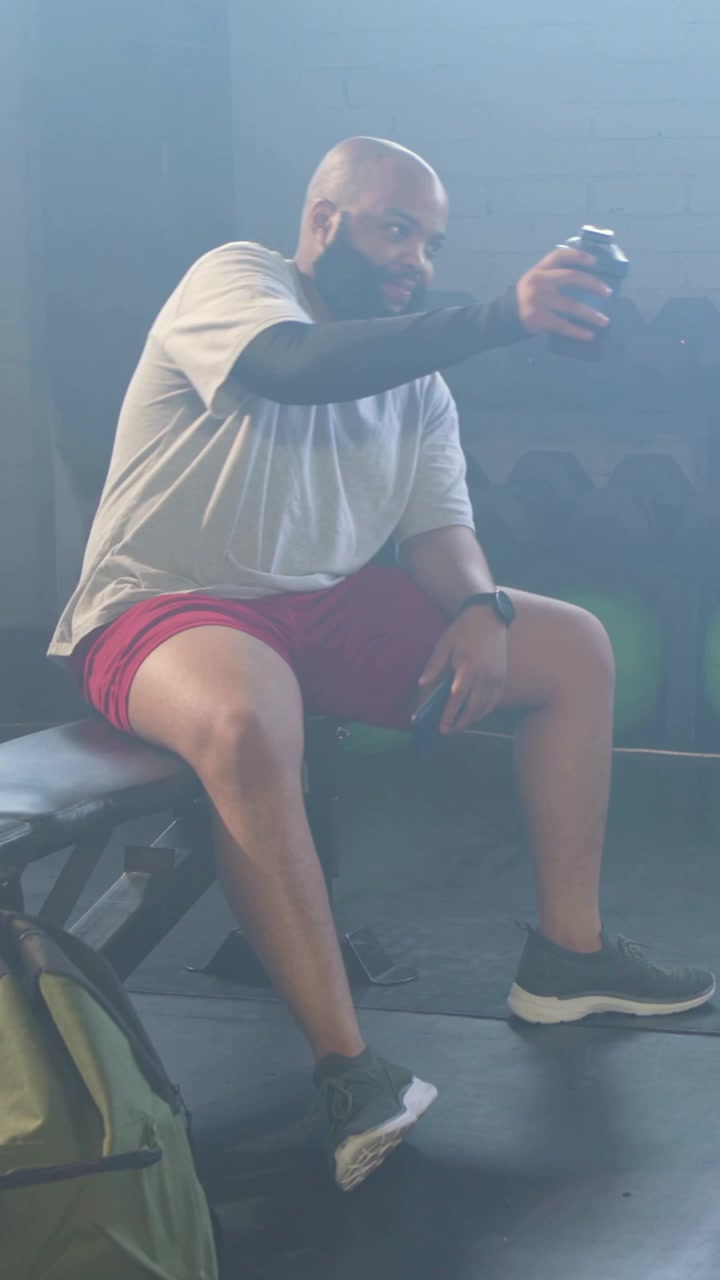 Video vertikal: Pria berlengan minum, duduk di bangku gym di antara set latihan, memegang shaker gelap