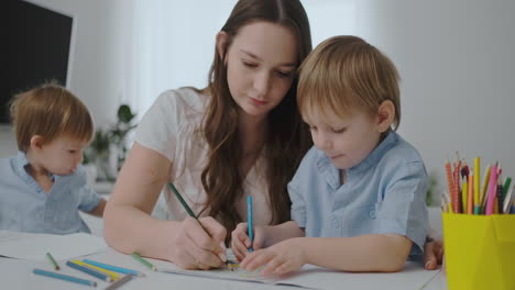 A-young-mother-with-two-children-sitting-at-a-white-table-draws-colored-pencils-on-paper-helping-to-do-homework