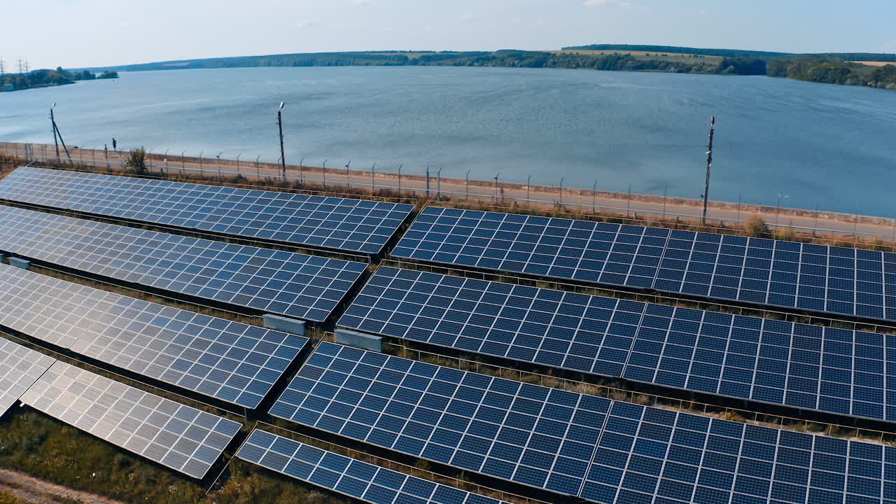Solar energy farm at sunlight. Photovoltaic sunny batteries near the river on blue water background. Alternative source of ecological energy. Camera rising.