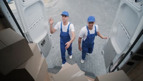 two young workers of removal company unload boxes and furniture from minibus into customer's home