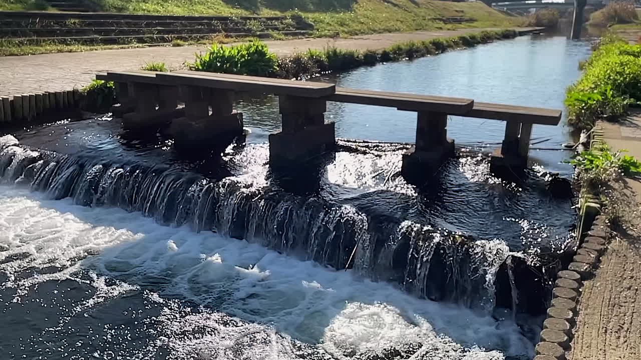 Slow motion drainage canal water flows over small pourover, sunny day